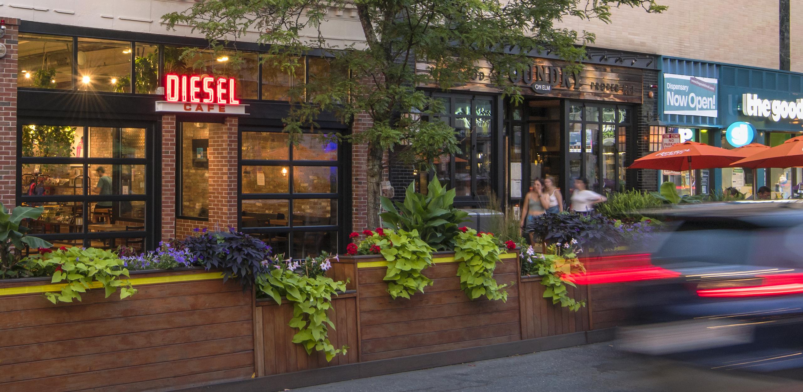 Diesel Café and adjacent shopfronts with wooden bank of seating and planters between sidewalk and roadway.