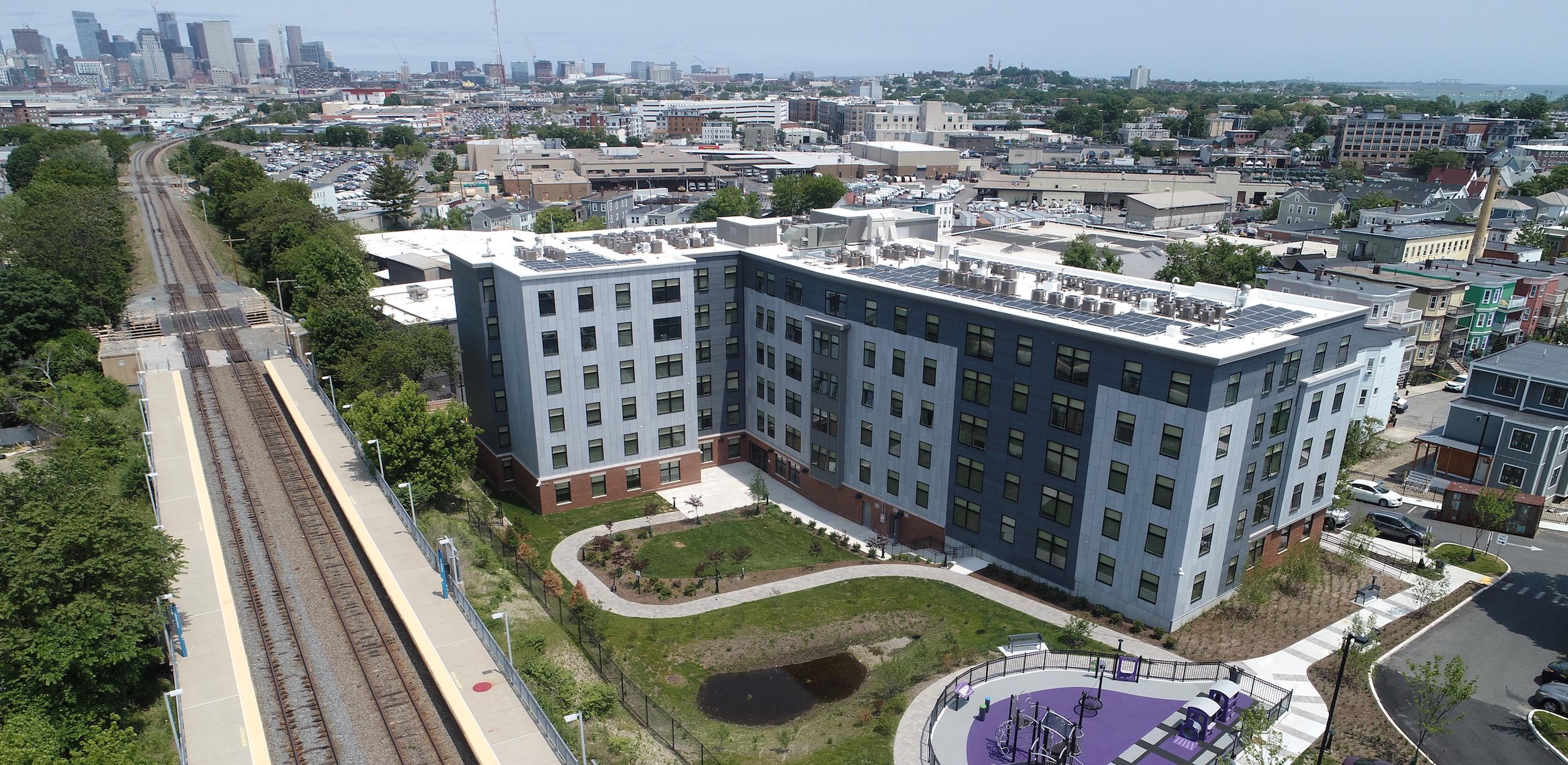 Bird's-eye view of residential building, playground and landscaping by adjacent commuter rail tracks.